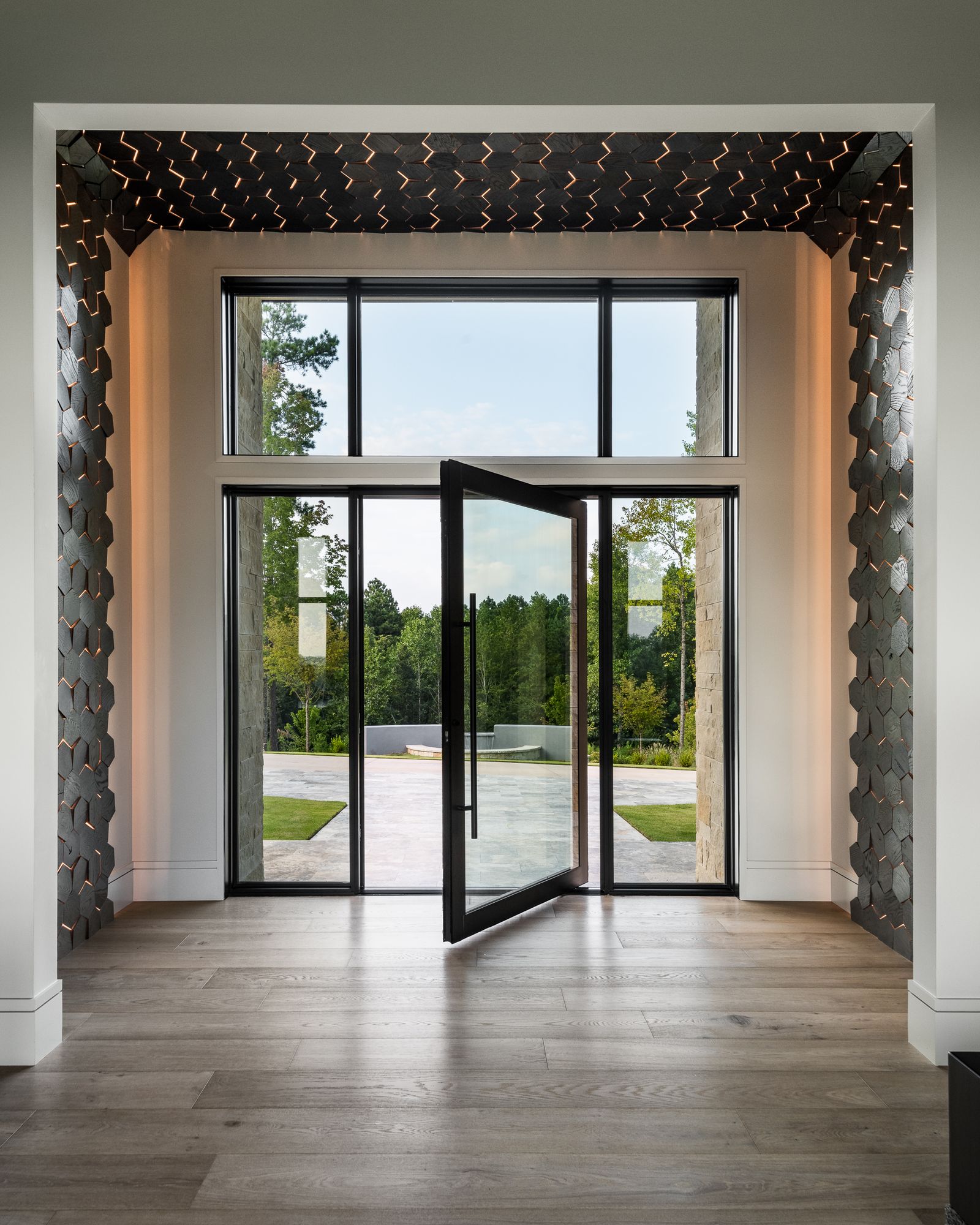 Dramatic luxury entryway with sculptural tile feature wall, pivot glass door, and wooded views — new construction interior design by Mac & Morgan Interiors, Upstate SC